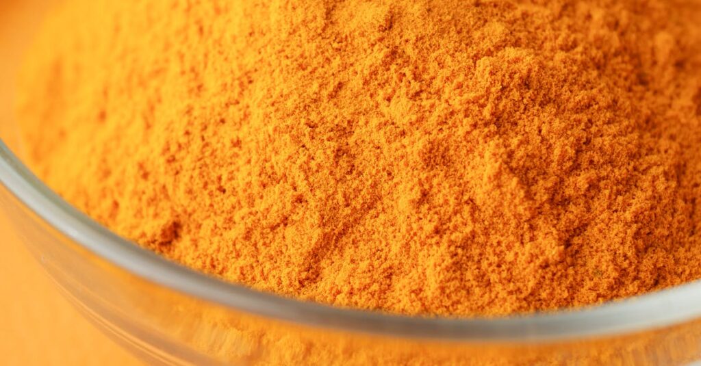 Close-up of bright turmeric powder in a glass bowl on an orange background, highlighting vibrant color contrast.