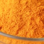 Close-up of bright turmeric powder in a glass bowl on an orange background, highlighting vibrant color contrast.