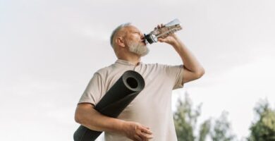Elderly man drinking water while holding a yoga mat outdoors, promoting fitness and hydration.